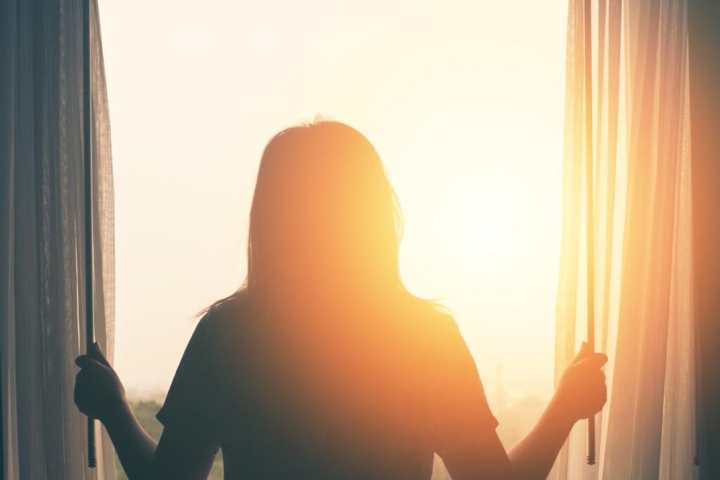 woman looking out window into the dawn sun practicing coping skills for anxiety that you should know about