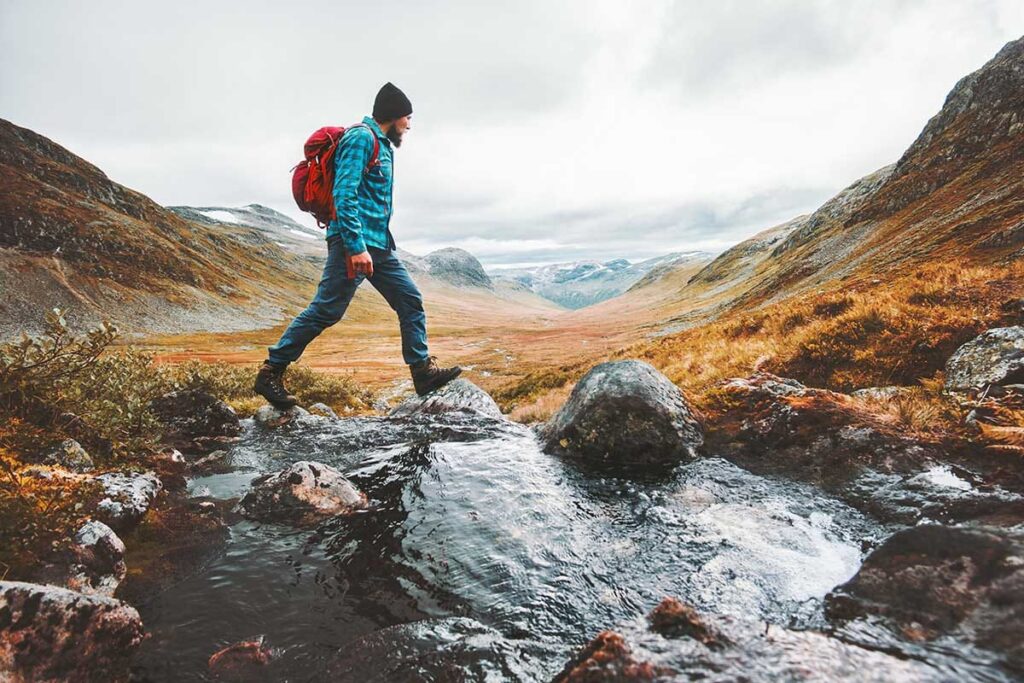 a person in a hat and backpack hikes through a stream in the mountains while wondering what is recreational therapy
