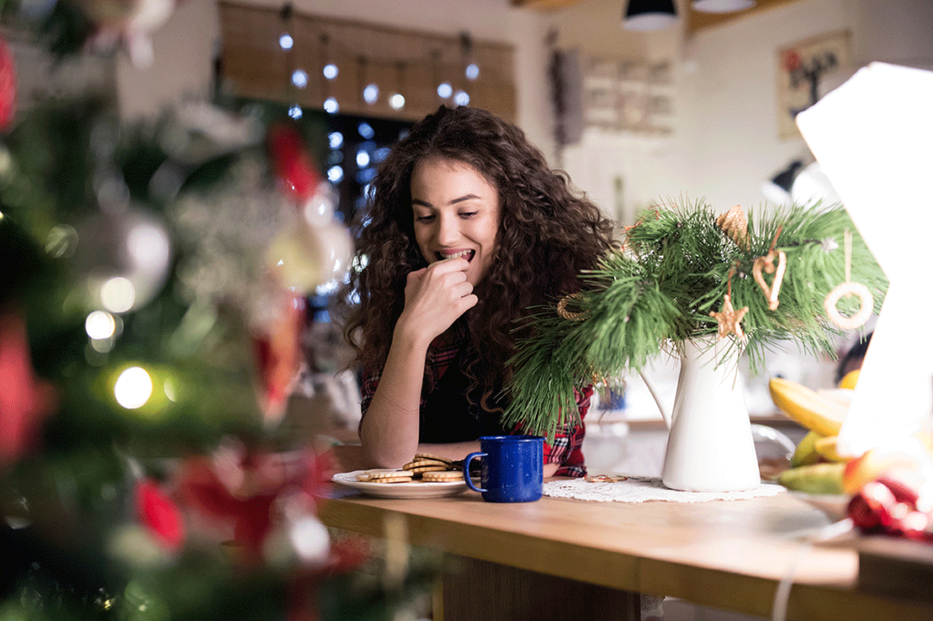 A woman eats cookies in front of the Christmas tree while she takes a break from her family. She's navigating holiday triggers.